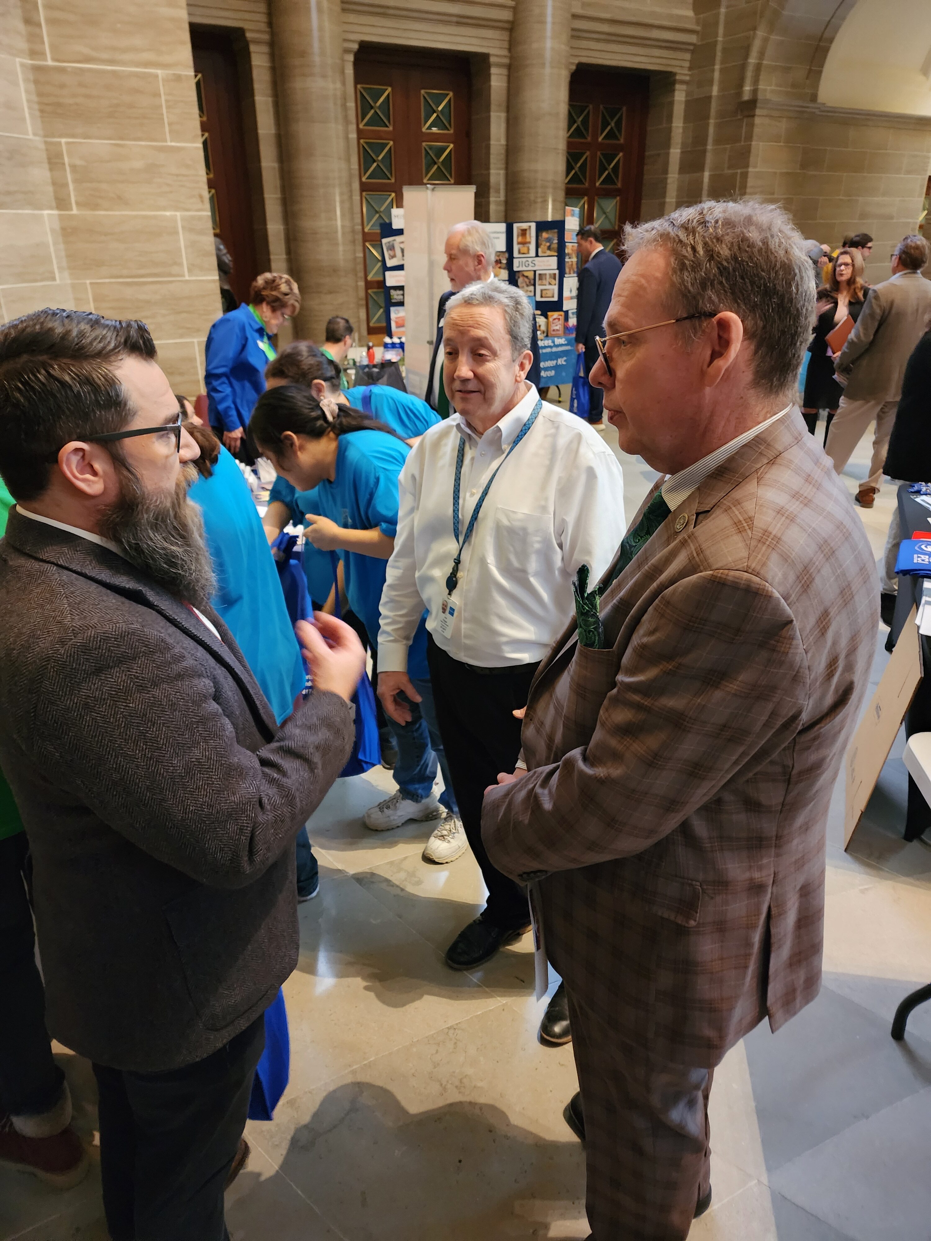 In the photo, Project CU Executive Director Kit Brewer stands on the far right wearing a plaid suit, while Project CU Sales Manager Jon Edgar, dressed in a white button-up shirt with a lanyard, is in the middle. They are engaged in conversation with Missouri State Representative Michael Burton on the far left, wearing a dark blazer. In the background, other people are interacting with various booths or displays at disability rights advocacy event in Jefferson City, Missouri in January 2024.