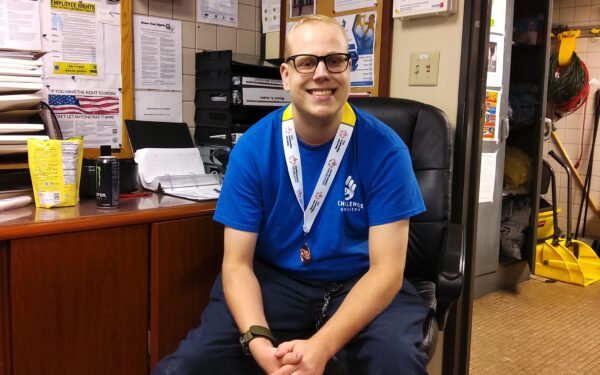 A man wearing glasses and a blue Challenge Unlimited t-shirt sits in an office, smiling at the camera. He is wearing a Special Olympics medal around his neck and has folded hands resting on his lap. The background includes a desk, bulletin boards, and a mop in a utility closet.