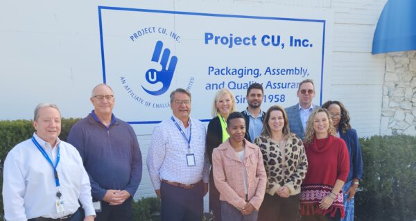 A group of nine people, including Project CU board members and staff, stand in front of a Project CU, Inc. sign on an exterior wall. The sign reads: 'Project CU, Inc. Packaging, Assembly, and Quality Assurance Since 1958. An affiliate of Challenge Unlimited.' The group is smiling and facing the camera on a sunny day.