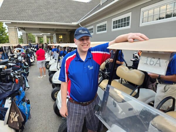 A man in a blue and red All-Star Training Club polo shirt and a matching hat is standing beside a row of golf carts at a golf event. He is smiling and resting his hand on cart number 16A, with other participants and carts in the background.