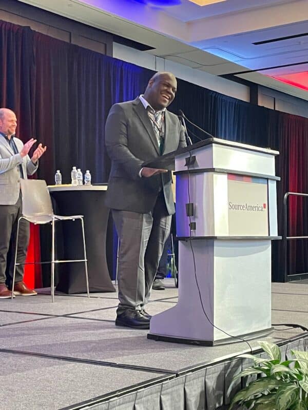 Jeffie Walker stands at a podium speaking confidently at the SourceAmerica Grassroots Advocacy Conference. He is smiling, dressed in a gray suit, and appears to be mid-speech. Behind him, a person is clapping, and the backdrop features black curtains with red and blue lighting. The SourceAmerica logo is visible on the podium.