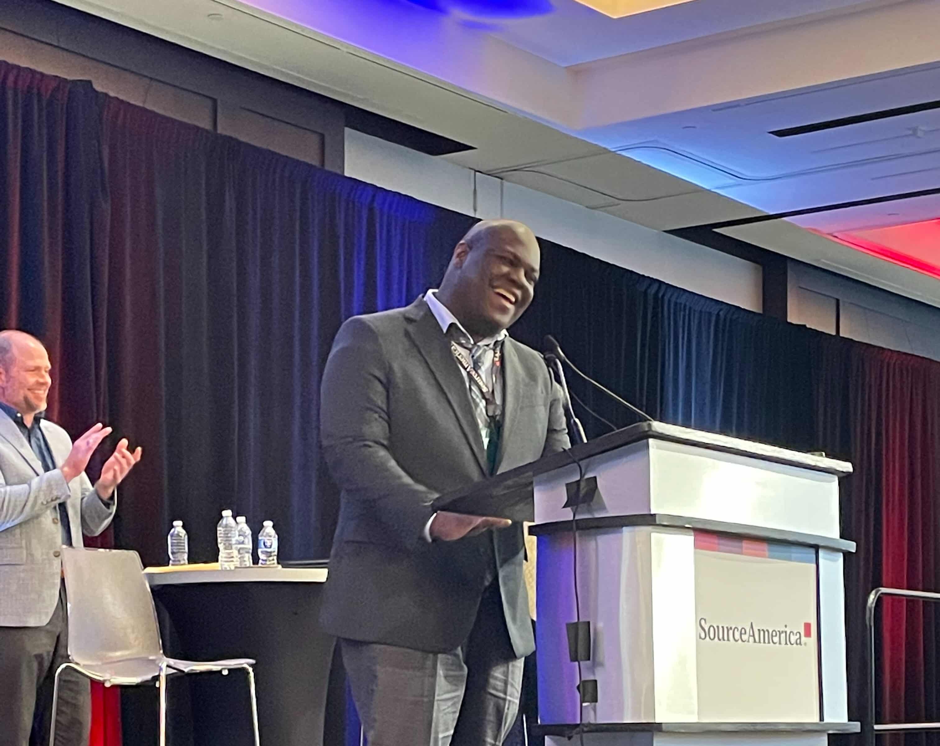 Jeffie Walker stands at a podium speaking confidently at the SourceAmerica Grassroots Advocacy Conference. He is smiling, dressed in a gray suit, and appears to be mid-speech. Behind him, a person is clapping, and the backdrop features black curtains with red and blue lighting. The SourceAmerica logo is visible on the podium.