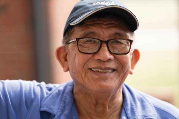 A smiling man wearing glasses and a dark blue baseball cap is looking directly at the camera. He is dressed in a light blue uniform shirt and standing outdoors with a soft blurred background.