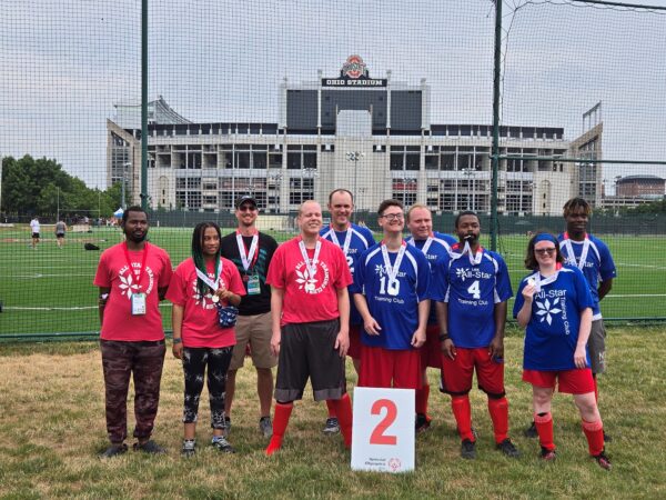 A group of nine individuals, five in blue jerseys and four in red t-shirts, pose together on a field in front of Ohio Stadium. They are part of a Special Olympics team and proudly display their medals. In front of the group is a 