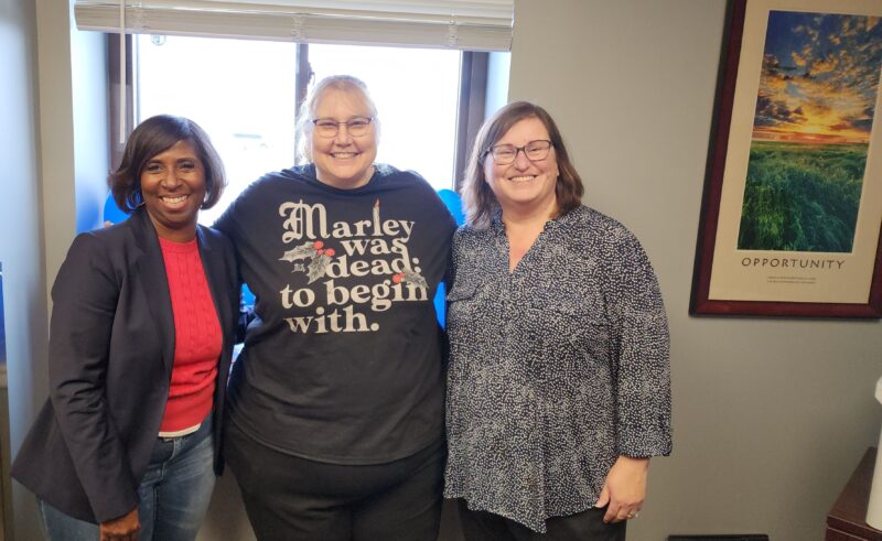 Charlotte Hammond, Janet Gentry, and Susan Wild smiling together in an office setting with a framed motivational poster reading "Opportunity" in the background.