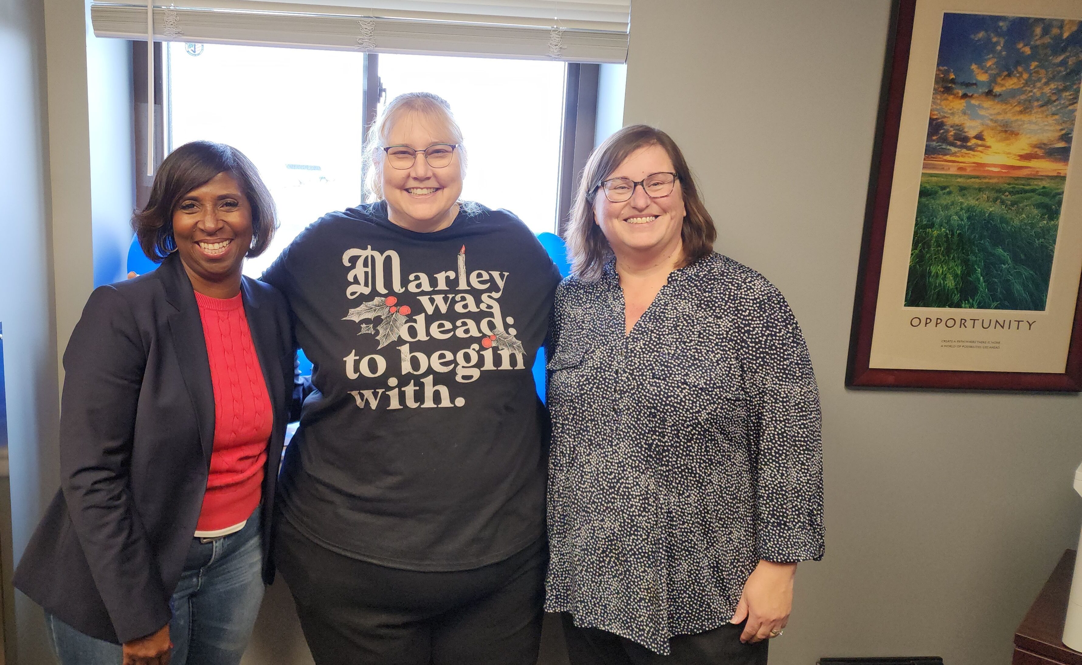 Charlotte Hammond, Janet Gentry, and Susan Wild smiling together in an office setting with a framed motivational poster reading "Opportunity" in the background.