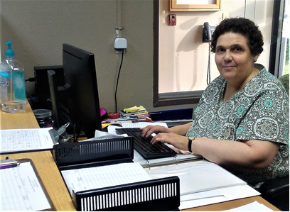 Stacey Springman sitting at her desk in the front office, working on a computer, surrounded by office supplies, showcasing her dedication and professionalism at Challenge Unlimited.